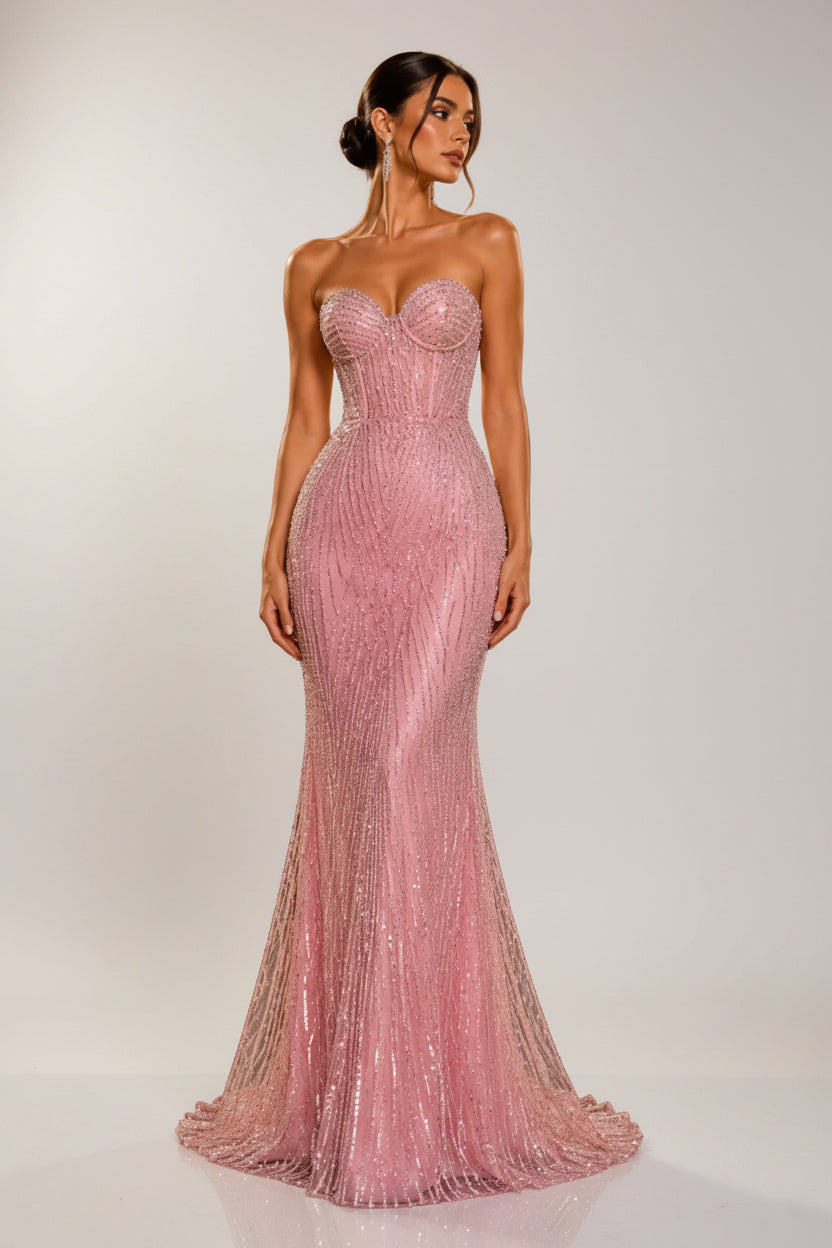 A woman models a strapless, sweetheart-neckline mermaid gown covered in sparkling pink sequins, set against a luxurious black marble and gold column background.