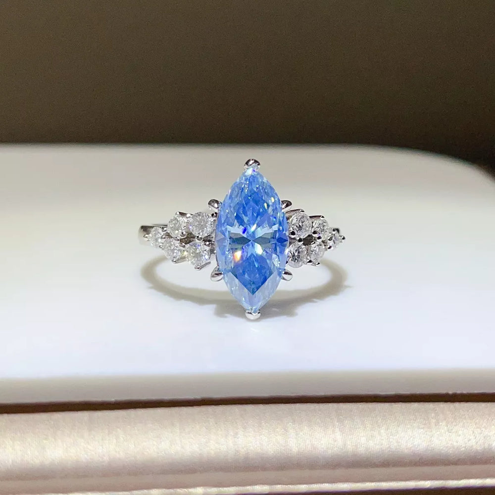 A silver ring featuring a marquise-cut light blue gemstone in the center, flanked by small clear stones on a white display surface.