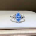 A silver ring featuring a marquise-cut light blue gemstone in the center, flanked by small clear stones on a white display surface.