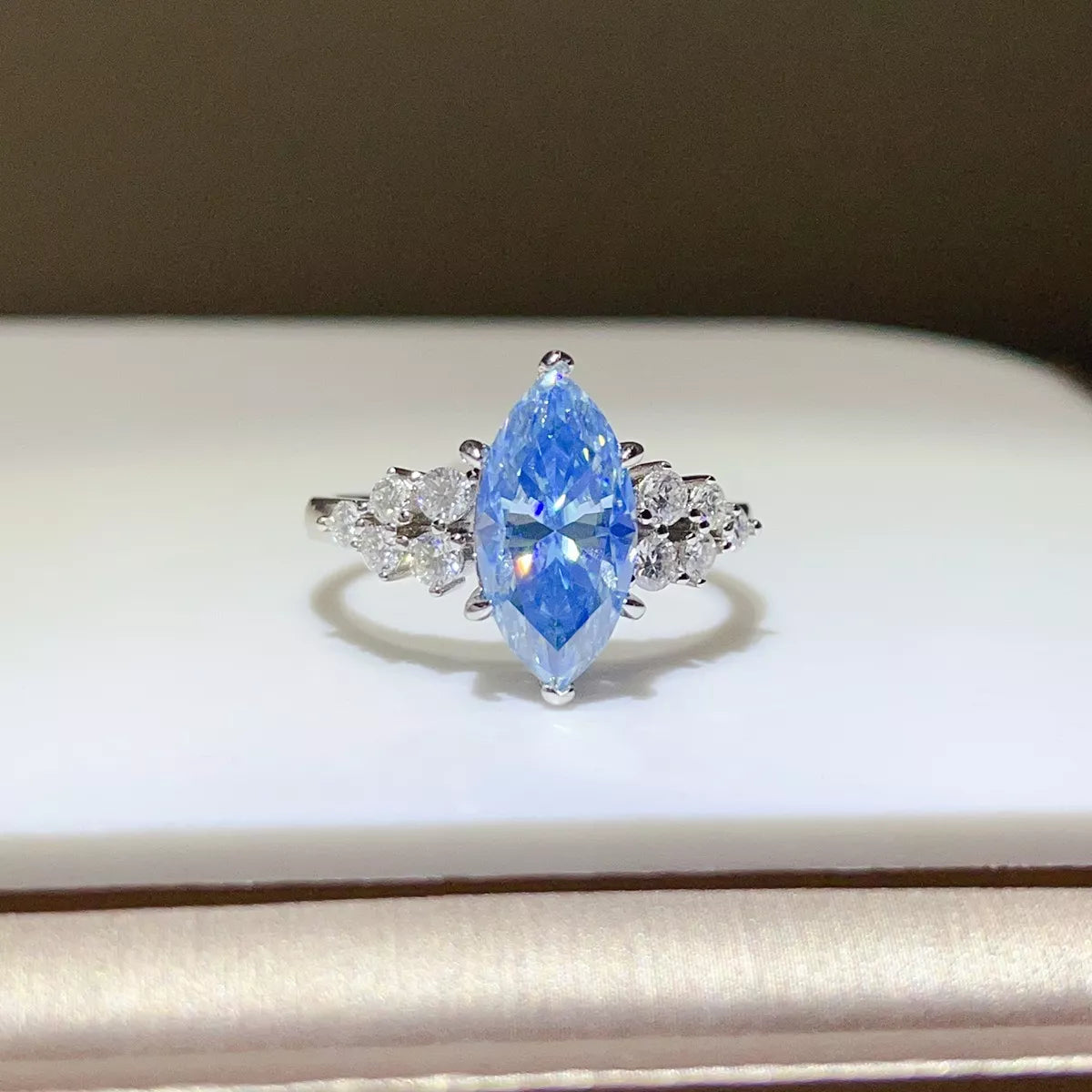 A silver ring featuring a marquise-cut light blue gemstone in the center, flanked by small clear stones on a white display surface.