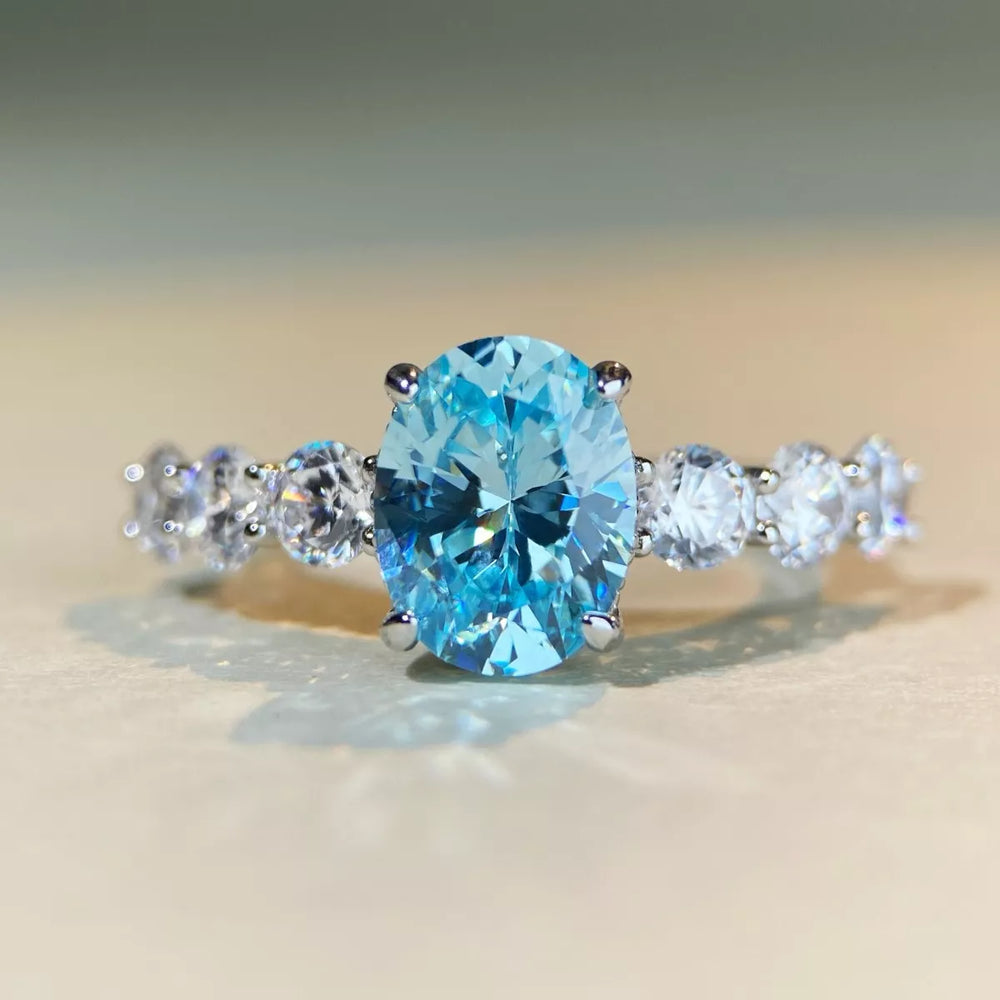 An oval-cut light blue gemstone ring set in a silver-colored band with three round clear accent stones on each side, photographed on a soft beige surface.