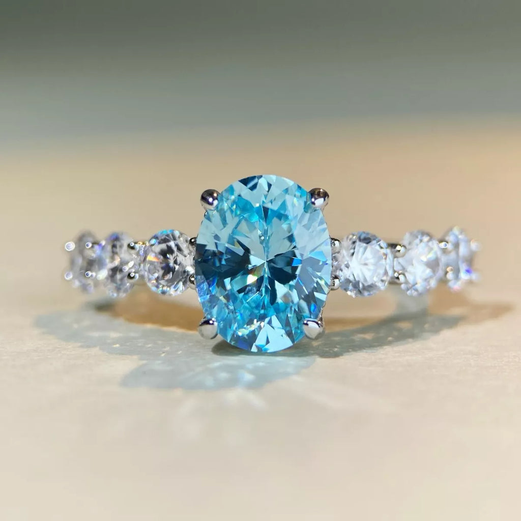 An oval-cut light blue gemstone ring set in a silver-colored band with three round clear accent stones on each side, photographed on a soft beige surface.