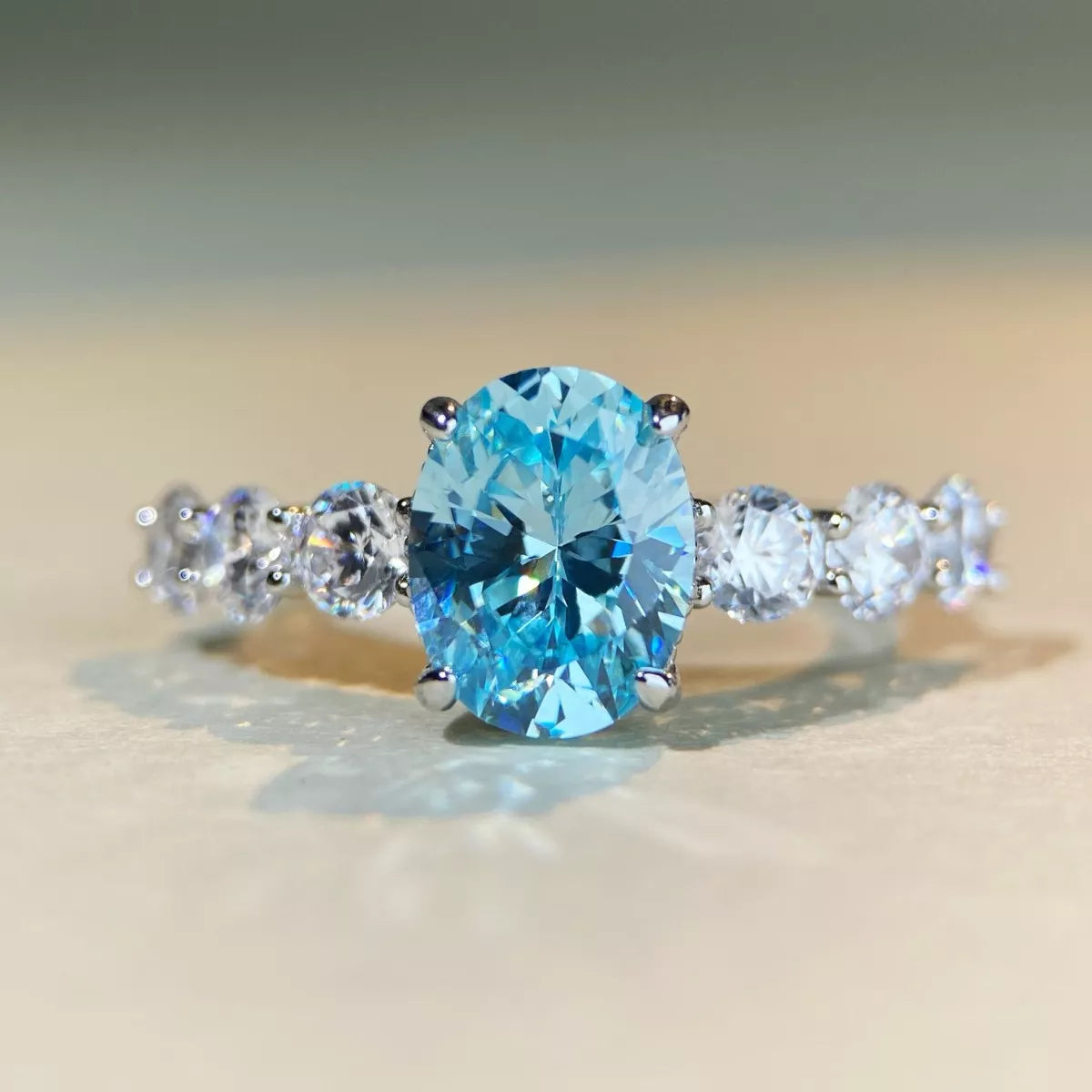An oval-cut light blue gemstone ring set in a silver-colored band with three round clear accent stones on each side, photographed on a soft beige surface.