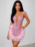 A woman modeling a light pink beaded mini dress with a plunging V-neckline, sheer mesh panels, feathered hem, and embellished shoulder details against a plain light gray wall.