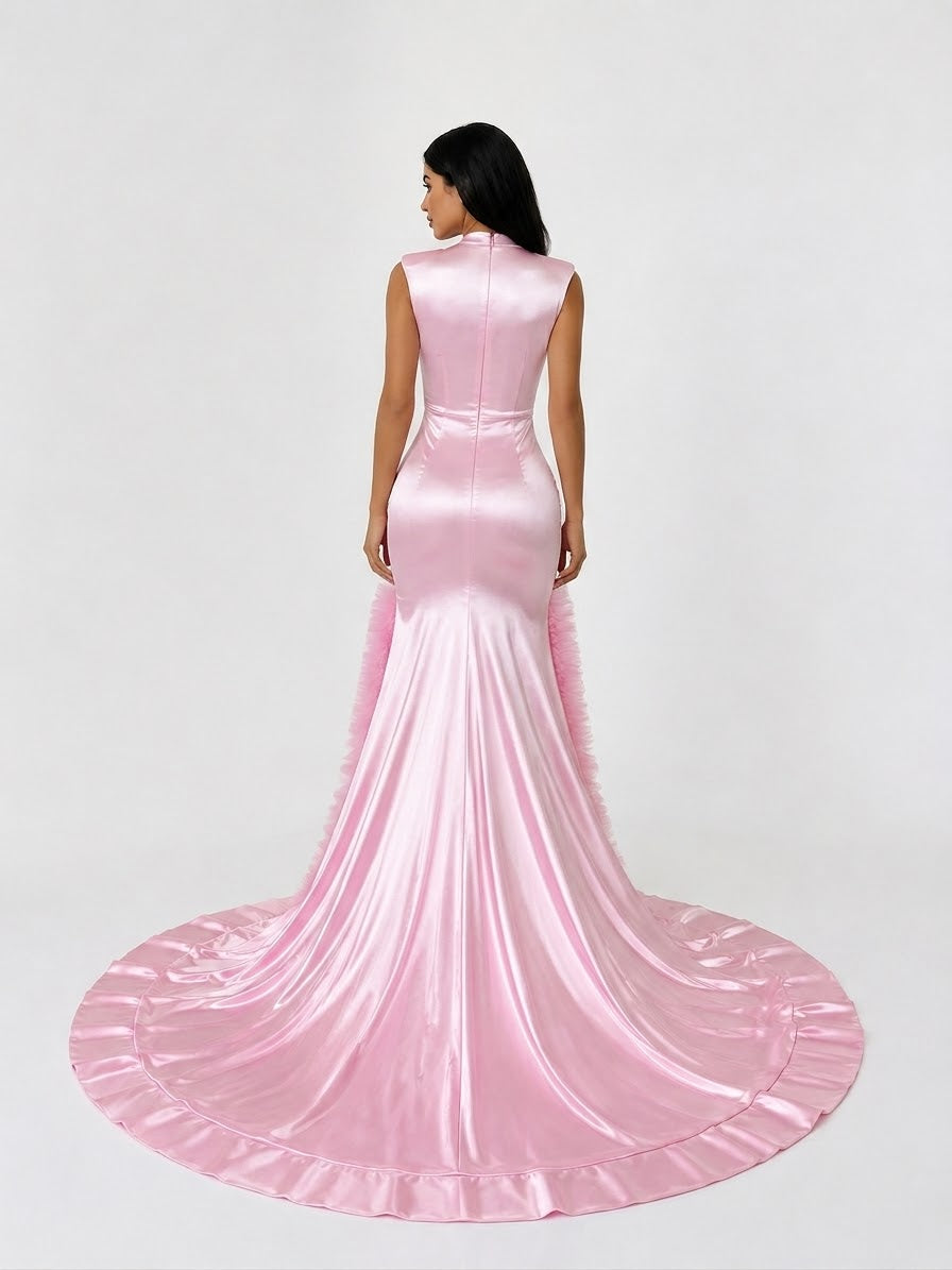 A woman modeling a sleeveless pink satin mermaid gown from the back, featuring a high neckline, fitted silhouette, and a long flared train on a light gray studio background.