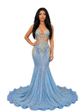 A woman models a floor-length light blue mermaid gown with gold sequin embroidery and a sheer lace bodice in an elegant indoor showroom. A mannequin wearing a champagne gown stands to the right, and a wall sign reading 'Confidential Couture' is visible behind her.