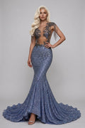 A blonde model wears a floor-length, mermaid-style evening gown with a sheer lace bodice, fringe details, and a sparkling blue sequined skirt with a slight train, posed against a neutral gray studio background.