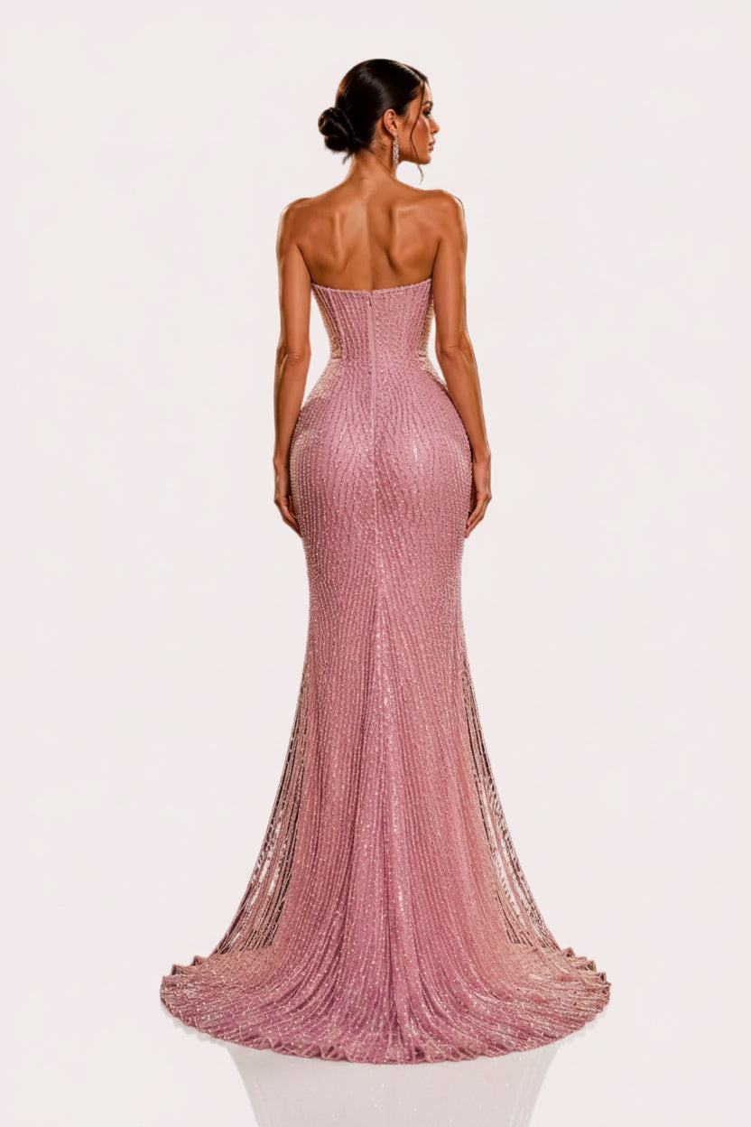 A woman stands with her back to the camera wearing a strapless, floor-length pink gown covered in shimmering sequins or beads. The setting is a dark, luxurious interior with gold marble walls, gold geometric decor, and warm spotlights.