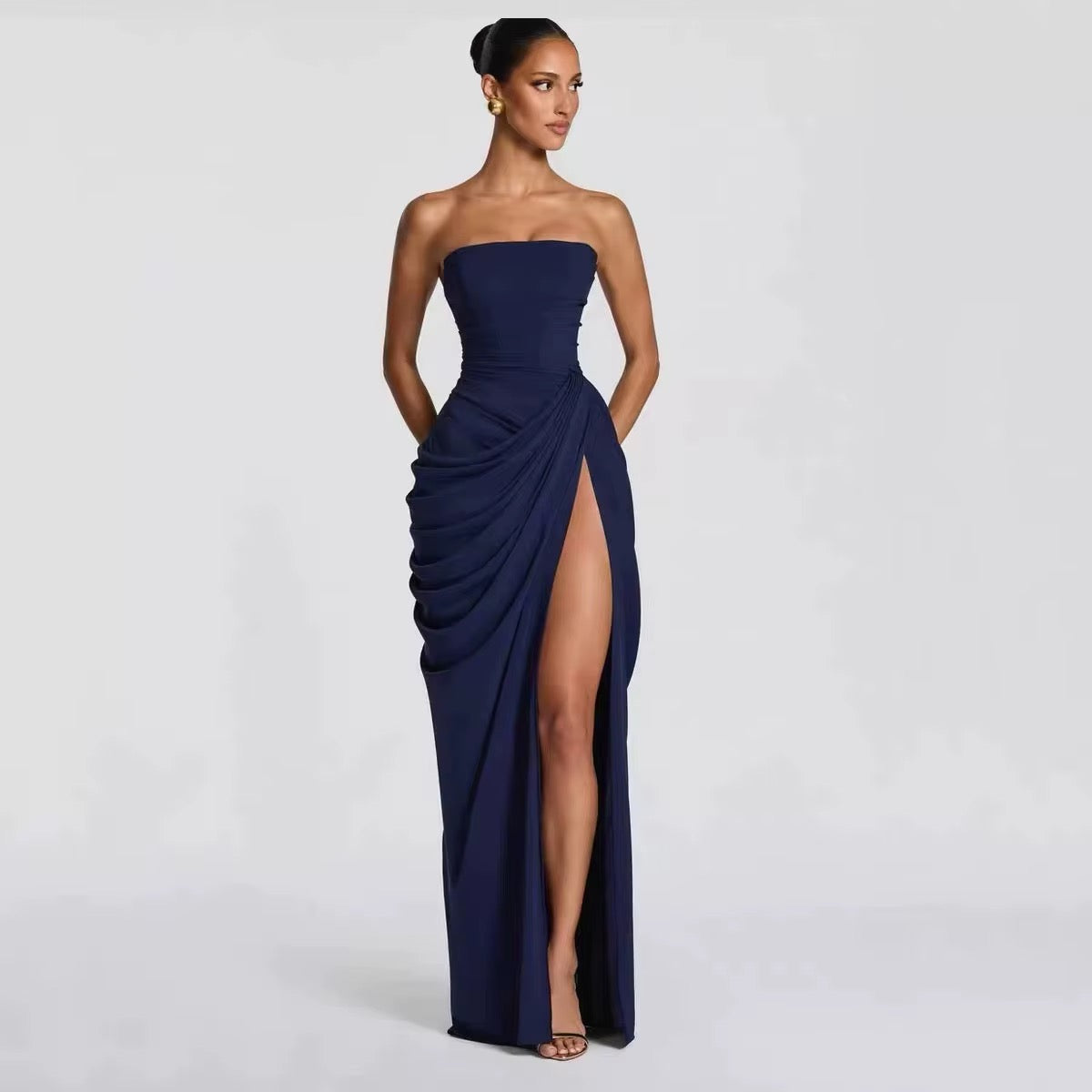 A woman modeling a strapless navy blue floor-length gown with a high thigh slit and draped gathered detailing on the skirt, photographed against a light gray studio background.