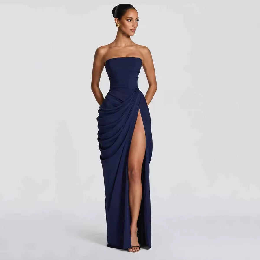 A woman modeling a strapless navy blue floor-length gown with a high thigh slit and draped gathered detailing on the skirt, photographed against a light gray studio background.