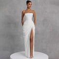 A woman models a strapless white floor-length gown with a ruched bodice, draped skirt, and high front slit, standing on a white round pedestal against a textured gray studio background.