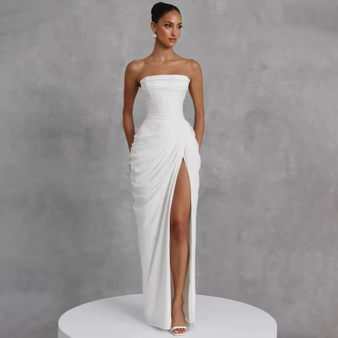 A woman models a strapless white floor-length gown with a ruched bodice, draped skirt, and high front slit, standing on a white round pedestal against a textured gray studio background.