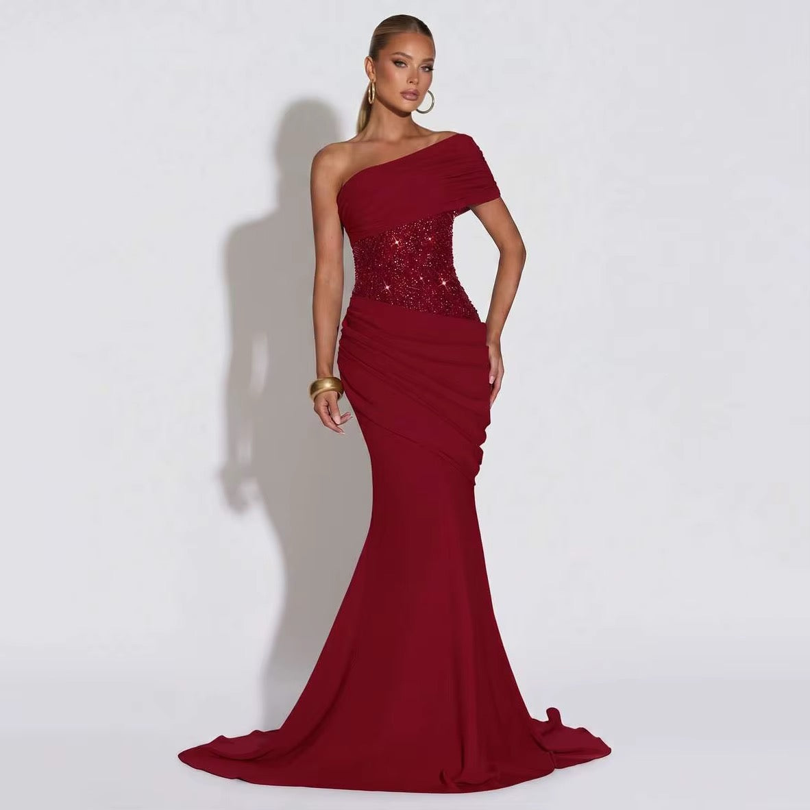 A woman models a floor-length, one-shoulder mermaid gown in deep burgundy with a heavily embellished sequined bodice, set against a plain light gray studio background.