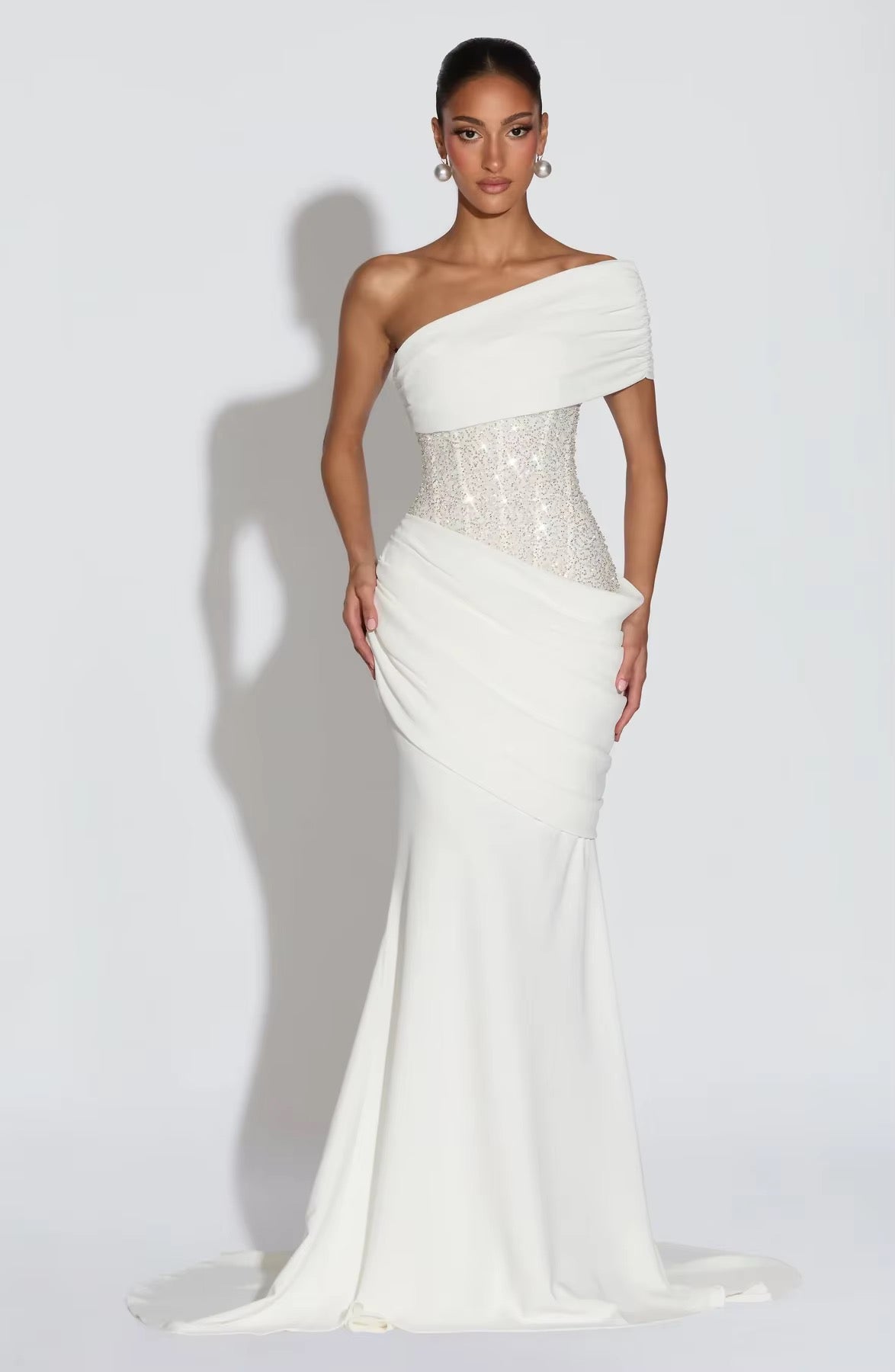 Woman wearing a one-shoulder white gown with a sequined bodice on a plain background