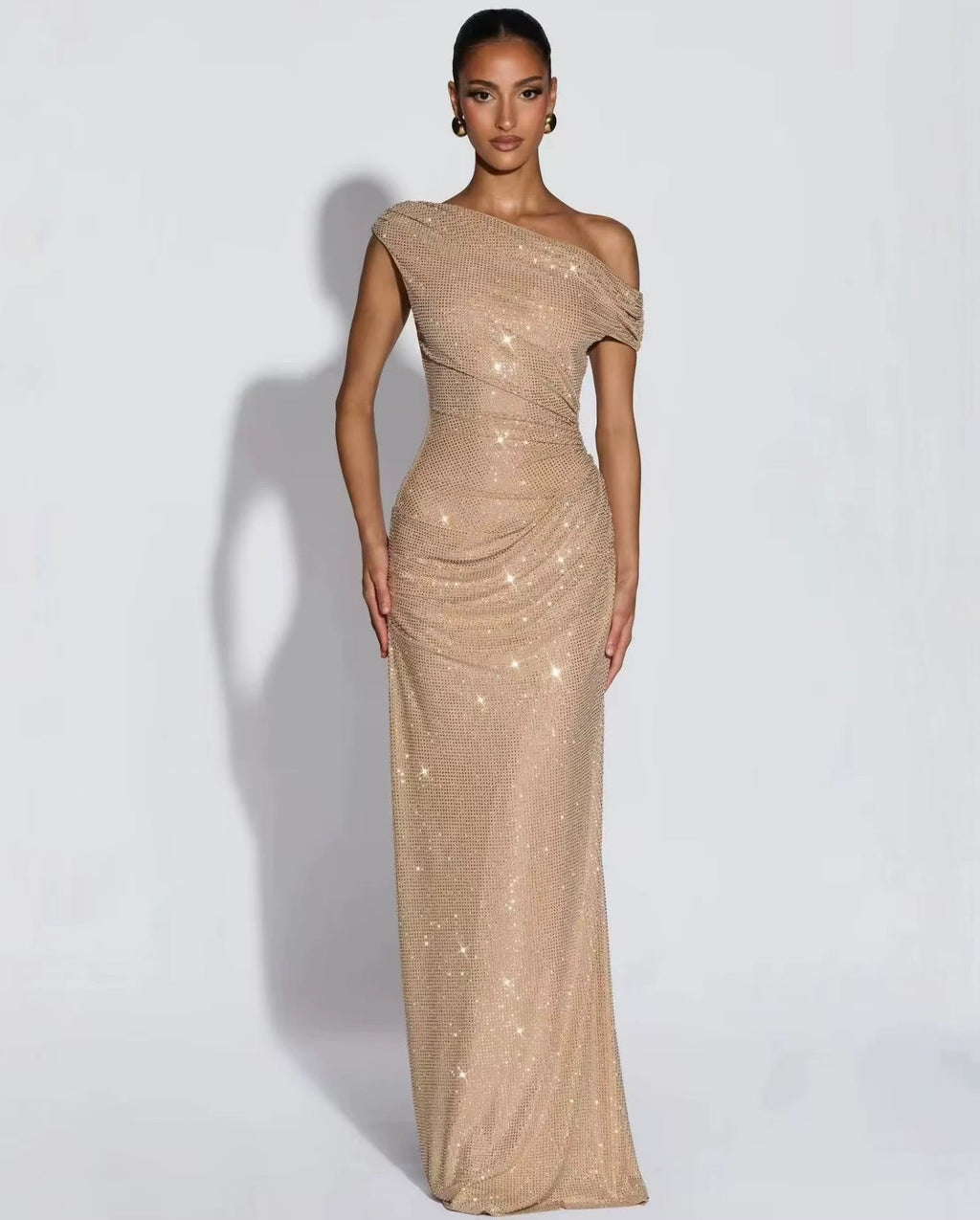 A woman modeling a floor-length, one-shoulder champagne sequin gown against a light gray studio background.