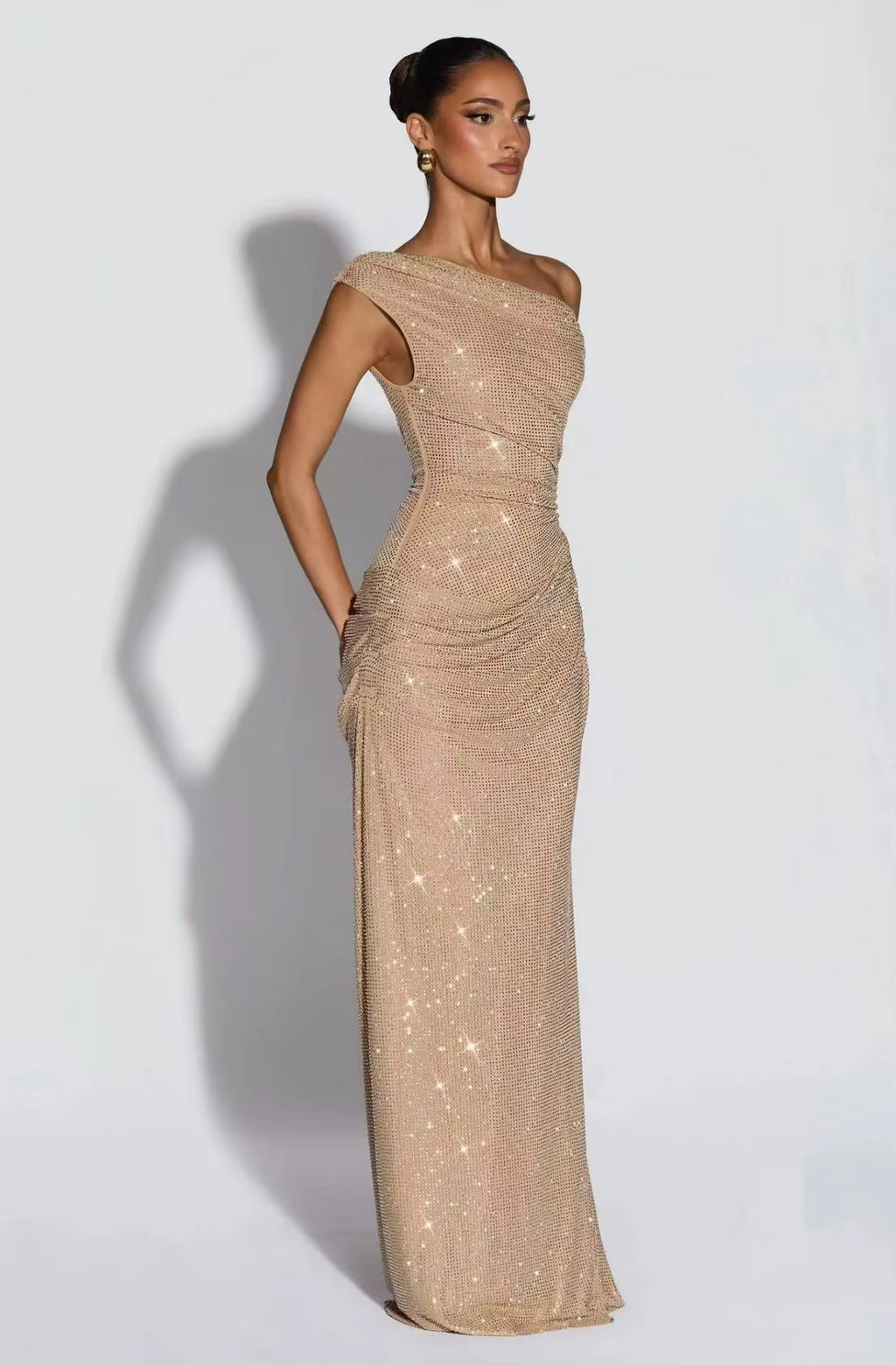 A woman modeling a floor-length, one-shoulder evening gown covered in shimmering sequins. The dress is a warm champagne or light gold tone and is photographed against a plain light gray studio background with a cast shadow.