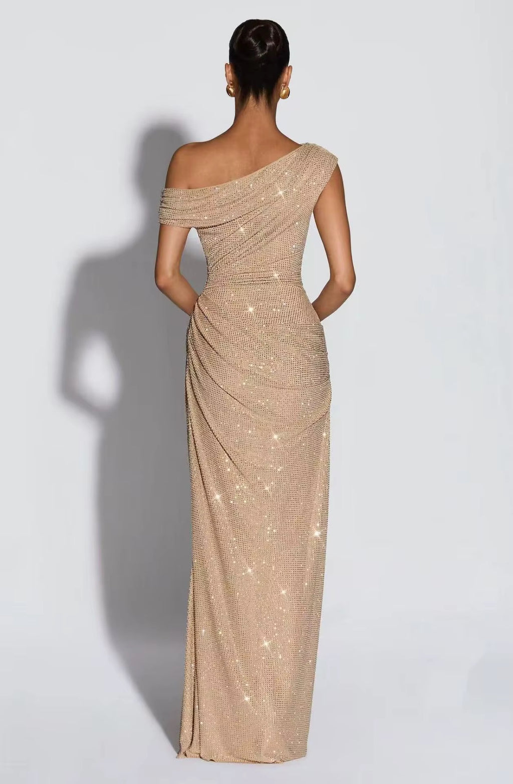 A woman viewed from the back wearing a floor-length one-shoulder evening gown covered in shimmering sequins. The dress is champagne beige with sparkling reflective details, photographed against a light gray studio background with a soft shadow cast to the left.