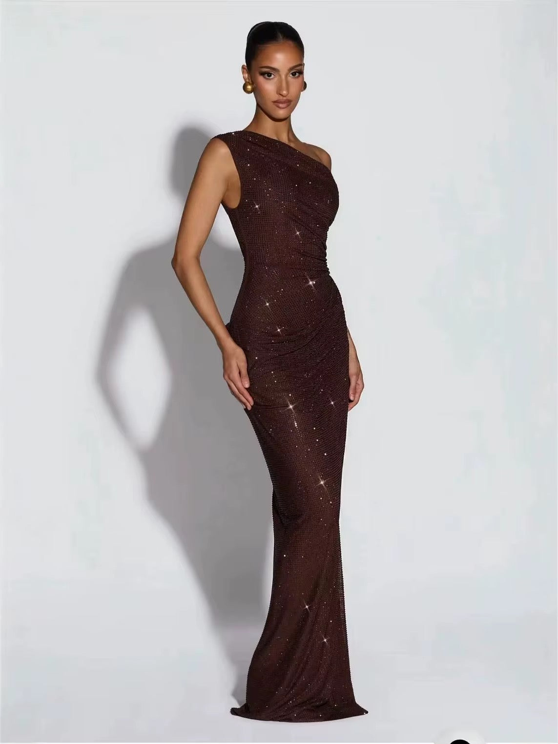 A woman modeling a floor-length, one-shoulder evening gown in dark brown with subtle shimmering embellishments, posed against a light gray studio background.