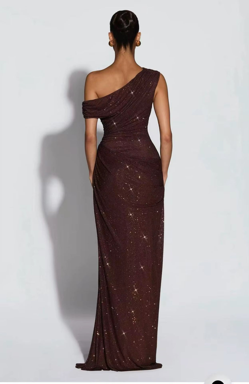 A woman stands with her back to the camera wearing a floor-length, one-shoulder evening gown in a deep burgundy-brown tone with scattered gold sequins, set against a plain light gray studio background.