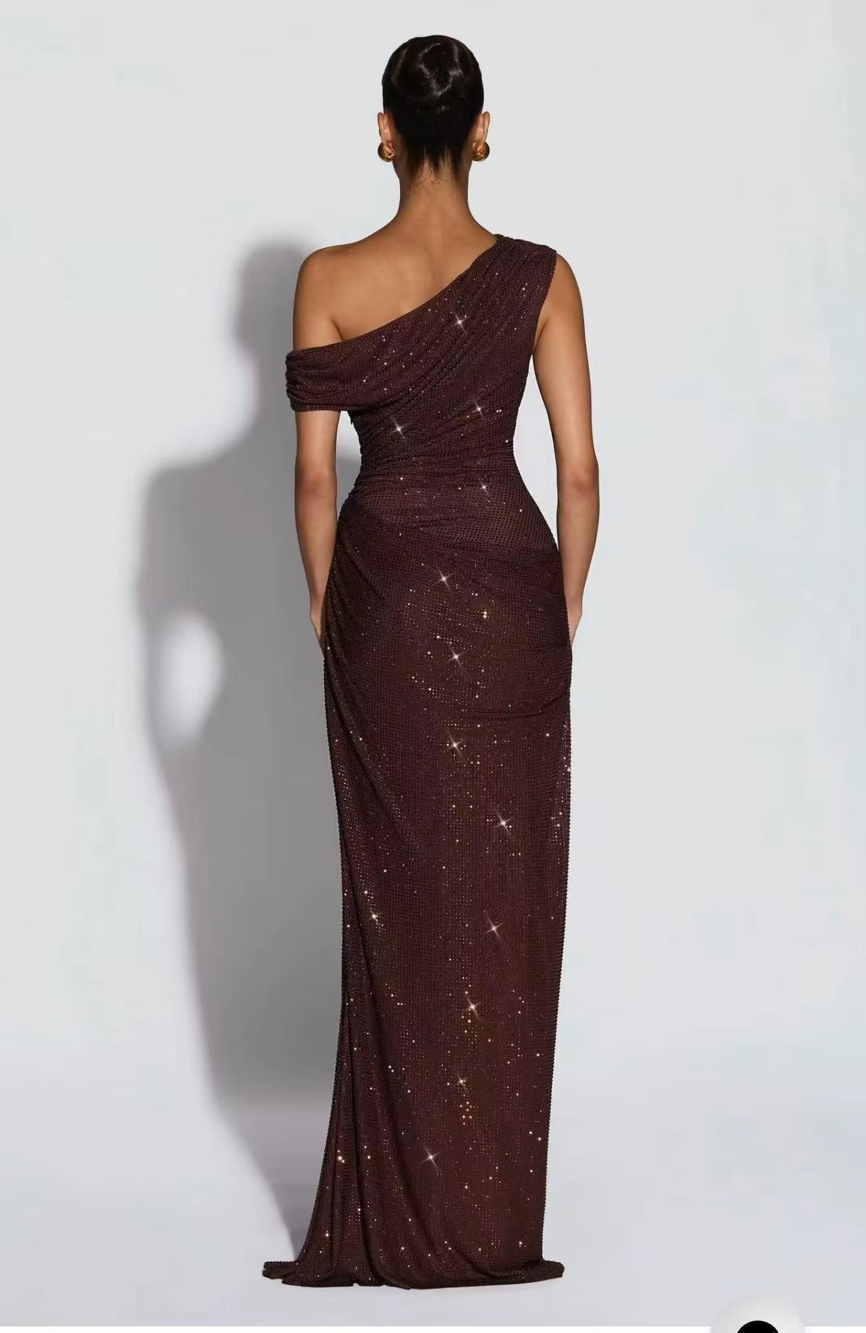 A woman stands with her back to the camera wearing a floor-length, one-shoulder evening gown in a deep burgundy-brown tone with scattered gold sequins, set against a plain light gray studio background.
