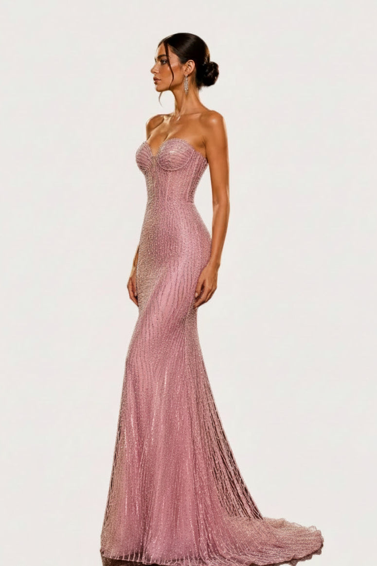 A woman modeling a strapless, floor-length pink gown covered in shimmering sequins and beads, standing in a luxurious black and gold interior with decorative circular lights and a marble pedestal.
