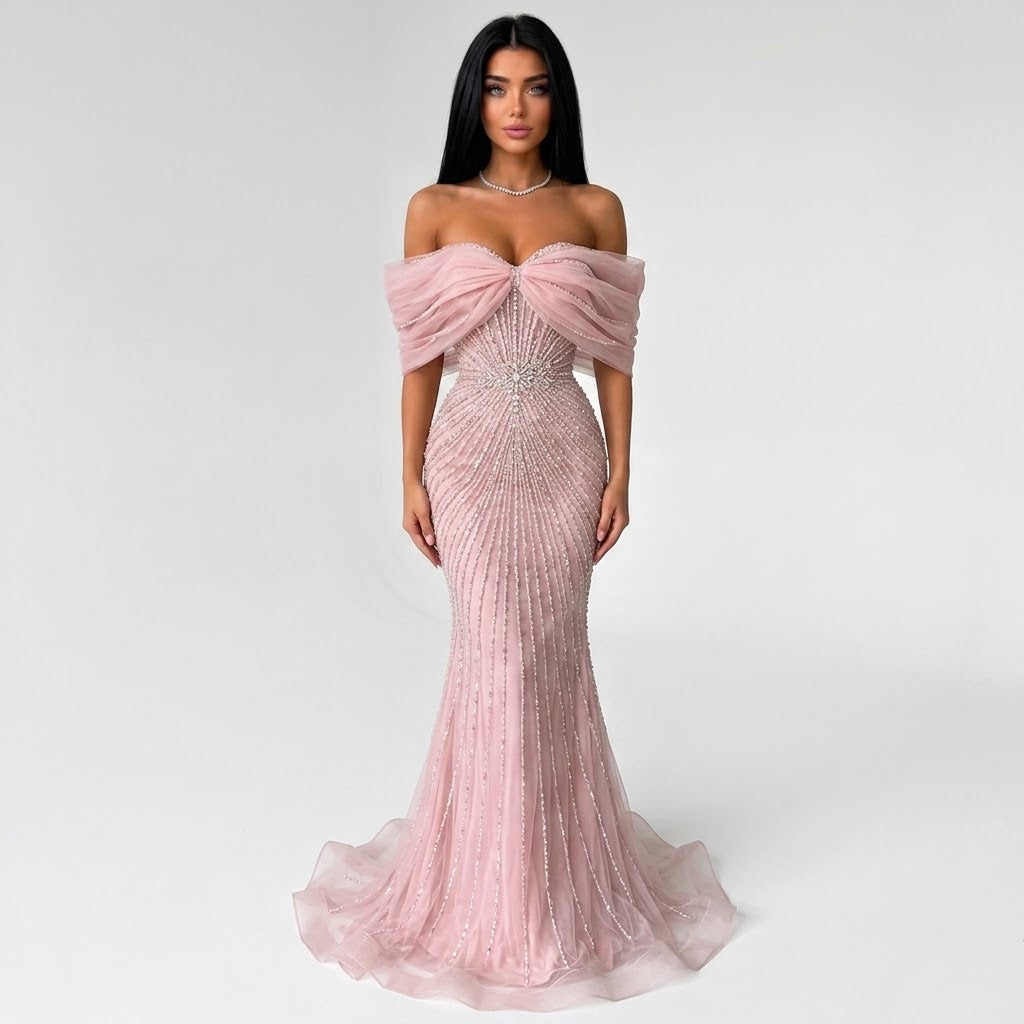 A woman modeling a floor-length off-the-shoulder pink gown with a sweetheart neckline, structured sheer shoulder fabric, a fitted bodice covered in beaded vertical lines, and a flared tulle skirt, photographed against a light gray studio background.