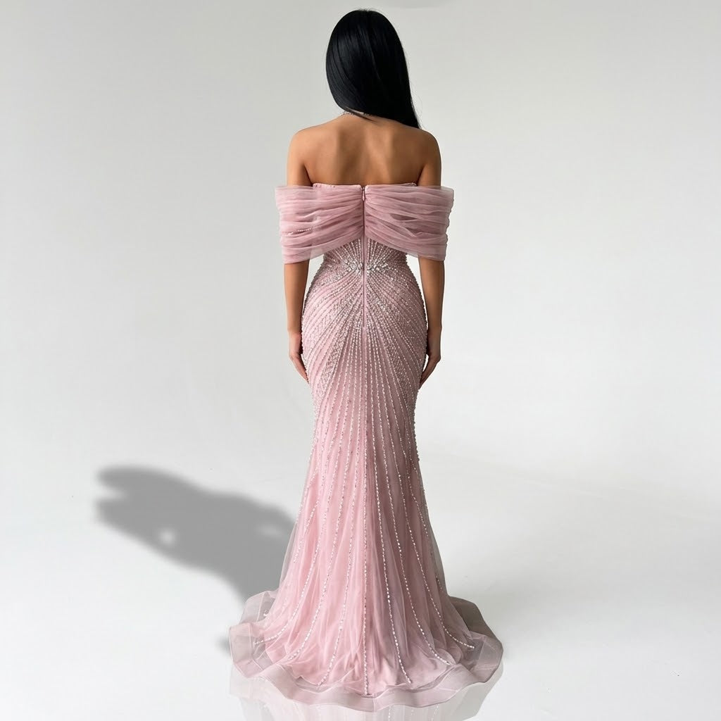 A woman stands with her back to the camera wearing an off-the-shoulder pink mermaid gown covered in sparkling beadwork and sequins, photographed against a plain light gray studio background.