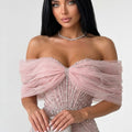 A woman wearing a pale pink off-the-shoulder evening gown with a large ruffled bow at the bust and dense bead embellishments, paired with a pearl necklace against a light gray studio background.
