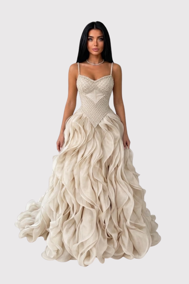 A woman modeling a floor-length champagne gown with a structured corset-style bodice, thin straps, and a voluminous layered ruffle skirt against a light gray studio background.