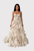 A woman modeling a floor-length champagne gown with a structured corset-style bodice, thin straps, and a voluminous layered ruffle skirt against a light gray studio background.