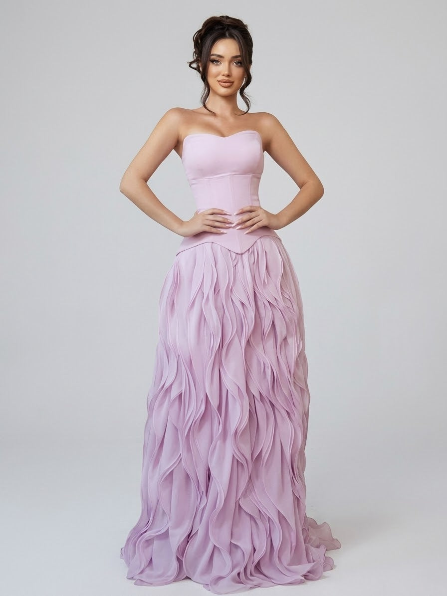 A woman modeling a strapless formal gown in a soft pastel lavender-pink color, featuring a structured corset-style bodice and a voluminous ruffled skirt, set against a plain light gray studio background. By KONFIDENTIAL KOUTURE