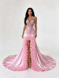 A woman modeling a formal pink gown with a deep V-neckline, heavily embellished bodice, thigh-high slit, and a dramatic ruffled train on a light gray studio background.