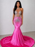 A woman modeling a bright pink mermaid gown with heavy silver rhinestone embellishments, a halter neckline, and a long ruffled train against a plain light gray wall.