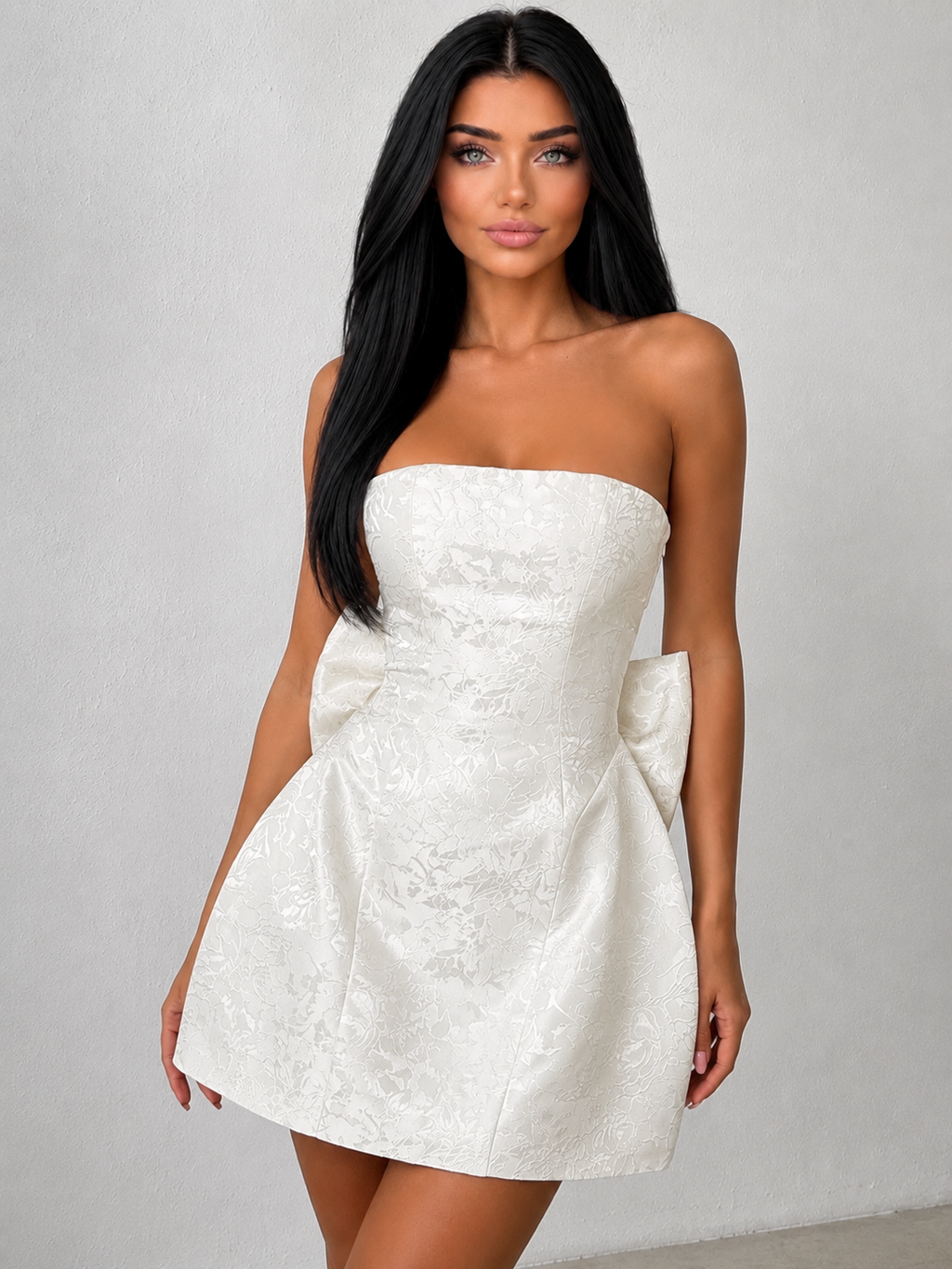 A woman modeling a strapless white mini dress with a textured jacquard pattern and large bow details at the waist, set against a neutral gray studio background.