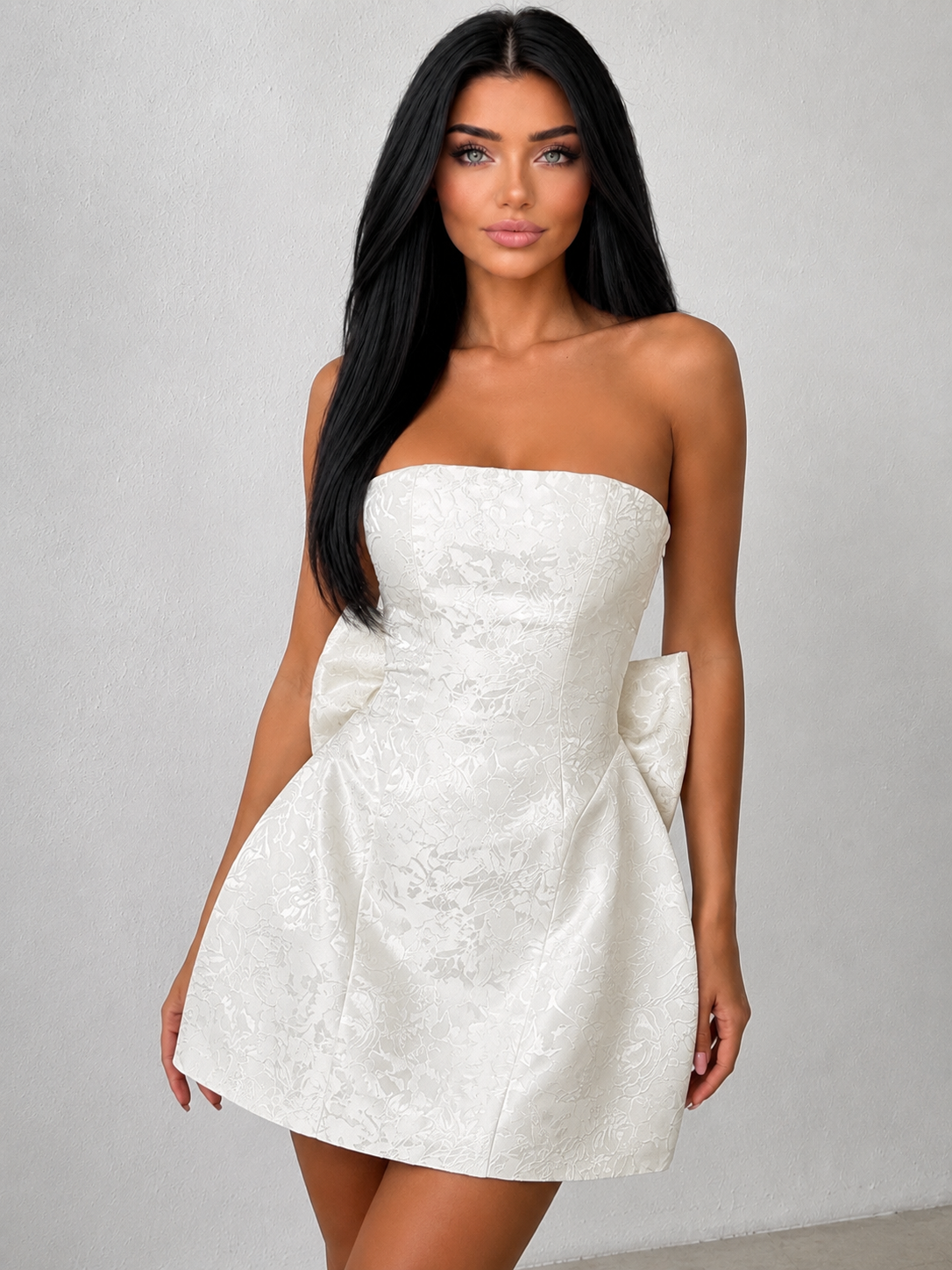 A woman modeling a strapless white mini dress with a textured jacquard pattern and large bow details at the waist, set against a neutral gray studio background.