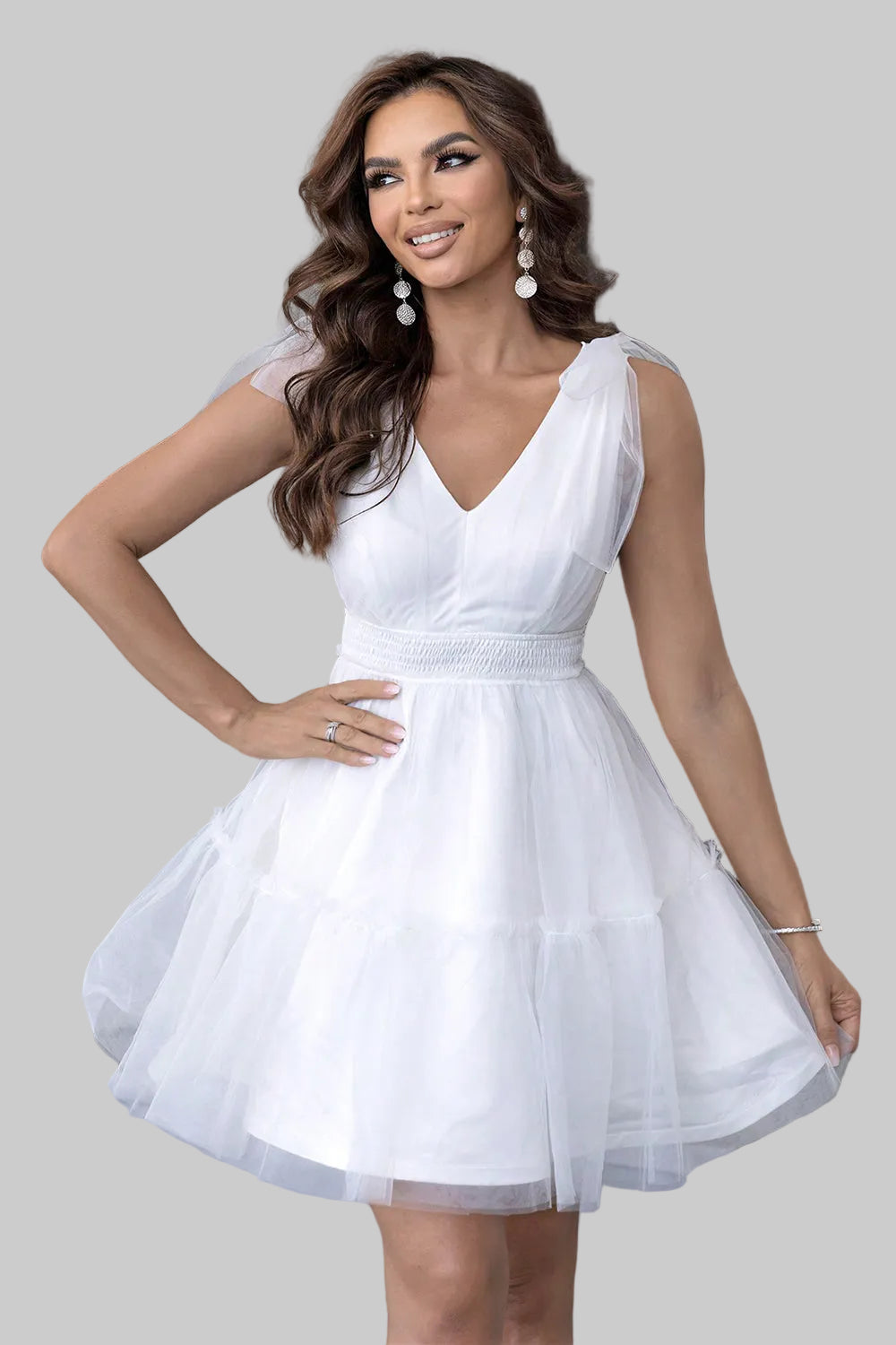A woman modeling a short white sleeveless dress with a V-neckline, fitted waist, and a flared tulle skirt, posed in front of a modern building entrance.