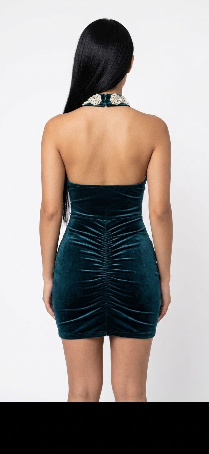 A woman shown from the back wearing a strapless dark teal velvet mini dress with ruching and a jeweled halter-style neckline, against a plain light background.