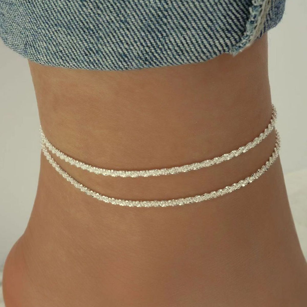 A close-up of a person's ankle wearing two delicate silver chain anklets, with the hem of blue denim jeans visible at the top.