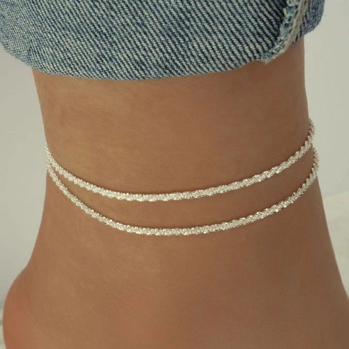 A close-up of a person's ankle wearing two delicate silver chain anklets, with the hem of blue denim jeans visible at the top.