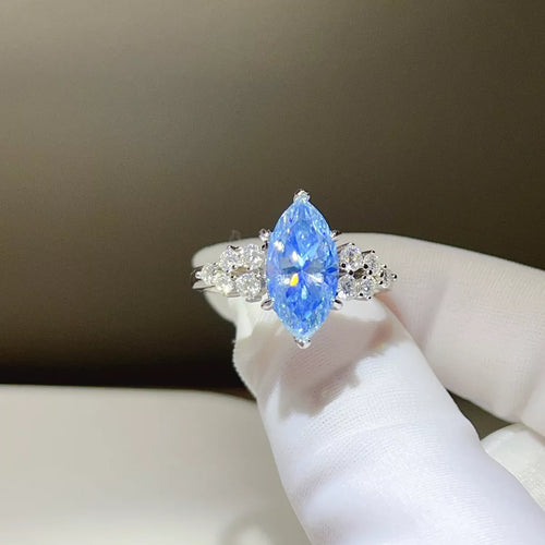 A silver ring featuring a marquise-cut blue gemstone at the center, flanked by small clear accent stones, held between white gloved fingers against a soft brown background.