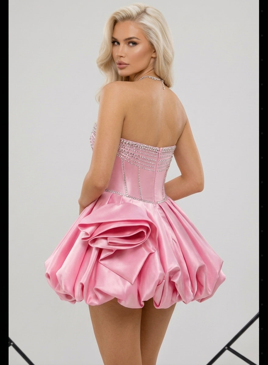 A blonde model shown from the back wearing a short pink strapless dress with a ruffled skirt and beaded corset-style bodice against a light gray studio background.