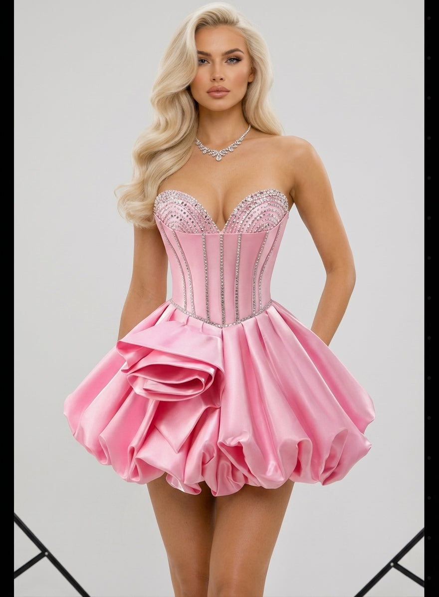 A blonde model wearing a pink strapless mini dress with a corset-style bodice, silver rhinestone trim, and a ruffled tiered skirt, photographed against a light gray studio background.