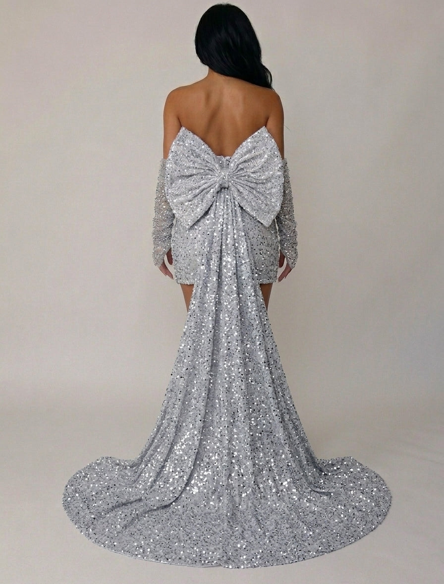 A woman shown from the back wearing a silver sequined off-the-shoulder gown with a large bow at the back of the neckline and a long train, photographed against a plain light beige studio background.