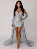 A woman modeling a silver sequined off-the-shoulder mini dress with long gloves and a long train, photographed against a plain light gray studio background.